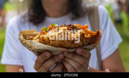 Frau hält halb gegessenes knuspriges Shawarma-Sandwich in Papierschale im Freien, Straßensnack, helles Tageslicht, Nahaufnahme. Stockfoto