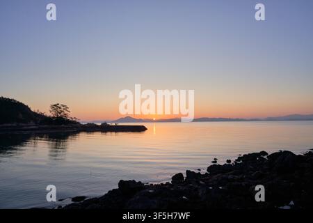 Sonnenaufgang über dem Meer in der Nähe von Sodeungseom, Südkorea, mit Menschen, die auf einer Mole als Silhouetten zu sehen sind, und fernen Bergen, die im Morgengrauen leuchten. Stockfoto