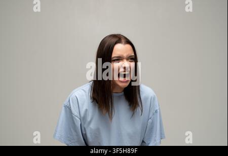 Junge Frau schreit aggressiv, drückt Wut und Frustration aus, schreit vor Wut auf grauem Hintergrund Stockfoto