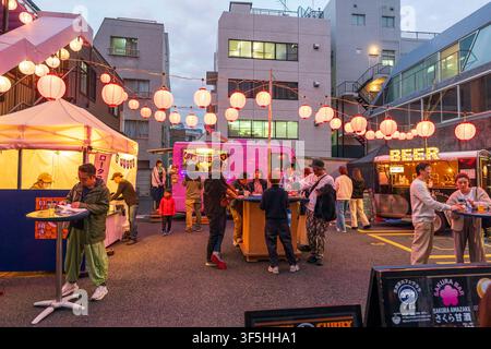 Tokio-Nachtfest mit Foodtrucks und Papierlaternen. Gruppen von Menschen und eine Familie genießen Essen und Getränke an Tischen, die auf einem Parkplatz aufgestellt sind. Stockfoto