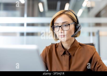Geschäftsfrau mit Headset und Brille, die an einem Laptop arbeitet und in einem modernen Büroumfeld engagierte Unterstützung und Kommunikation bietet Stockfoto