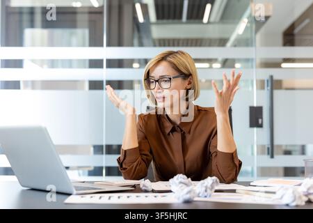 Junge Geschäftsfrau mit Brille, die Stress und Angst durch überwältigende Arbeitsprobleme erlebt, frustriert gestikulierend, während sie an einem Schreibtisch mit einem Laptop und zerknitterten Papieren sitzt Stockfoto