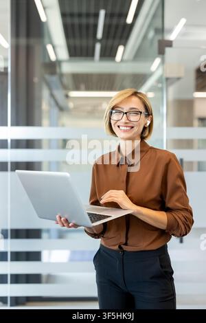 Professionelle Geschäftsfrau mit Brille und kabellosen Ohrstöpseln steht in einem zeitgenössischen Büro. Lächelnd selbstbewusst, während sie mit einem silbernen Laptop interagiert. Steht für Erfolg. Technologie Stockfoto