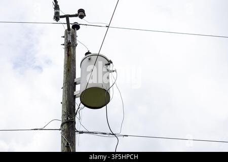 Elektrischer Verteilertransformator, der an einem Holzpfahl mit Stromleitungsanschlüssen montiert ist, vor einem Hintergrund aus bewölktem Himmel. Konzept der Stromversorgung. Stockfoto