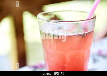 Nahaufnahme eines erfrischenden Glases hausgemachter Himbeerlimonade, serviert mit Eiswürfeln, frischen Minzblättern und einem rosa Trinkhalm auf einem Tisch. Stockfoto