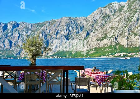 Eine malerische Außenterrasse eines Cafés am Meer mit Tischen mit karierten Tischdecken und Stühlen, vor dem atemberaubenden Hintergrund des blauen Meeres und massiver Felsberge. Stockfoto