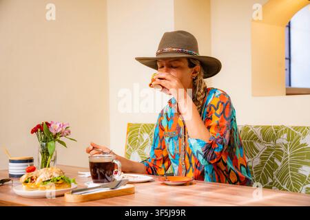 Stilvolle Frau mit breitkrempigem Hut trinkt Kaffee und genießt ein frisches Frühstückssandwich an einem hellen Cafétisch, umgeben von Blumen, Geschirr und Stockfoto