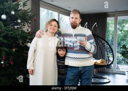 Lächelnde dreiköpfige Familie, die neben einem geschmückten Weihnachtsbaum in einem hellen modernen Wohnzimmer posiert, warmen Pullover und ein cremefarbenes Pullover-Kleid tragend; Vater h Stockfoto