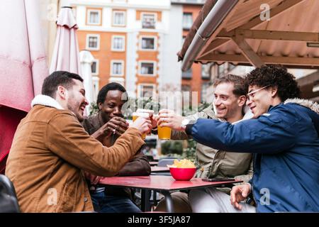 Eine Gruppe von Freunden genießt Getränke und Gelächter auf einer Barterrasse und prostet dem Glück inmitten einer lebendigen Stadtsilhouette zu. Diese Momente unterstreichen die Freuden von f Stockfoto