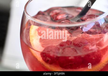 Eine detaillierte Nahaufnahme eines lebendigen rosa Gin-Cocktails, serviert in einem großen Glaskelch mit Eiswürfeln, frischen Himbeeren und einer Zitrusfrucht-Scheibe Stockfoto
