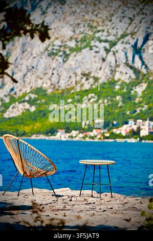 Eine ruhige, malerische Meeresansicht mit einem einzelnen geflochtenen Rattanstuhl und einem kleinen Beistelltisch, die auf einem rauen Steinpier platziert sind, mit dem blauen Adriatischen Meer, einem Küstendorf und massiven Bergen im Hintergrund. Stockfoto