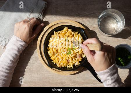 Ansicht von oben einer Person, die hausgemachten Mac und Käse aus einer schwarzen Pfanne isst, garniert mit Petersilie und serviert auf einem Holztisch neben Wasser und Kräutern Stockfoto