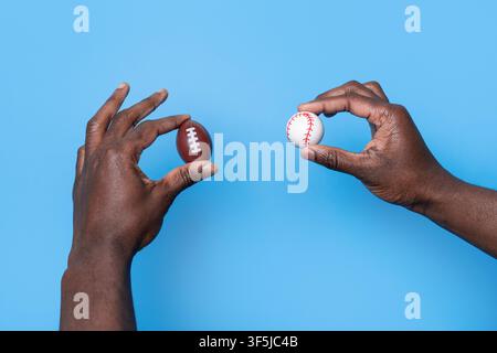 Zwei Hände halten Mini-Fußball- und Baseballbälle vor einem hellblauen Hintergrund und veranschaulichen den Kontrast zwischen beliebten amerikanischen Mannschaftssportarten Stockfoto