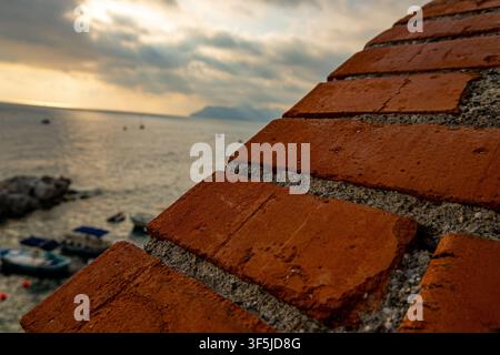 Nahaufnahme einer verwitterten roten Backsteinmauer mit Blick auf das Meer bei Sonnenuntergang Stockfoto