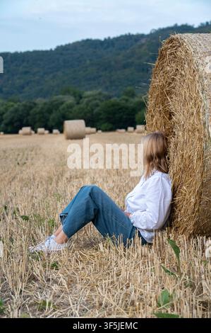 Unerkennbare Frau sitzt an einem runden Heuballen auf einem abgeernteten Feld mit ländlicher Landschaft Stockfoto
