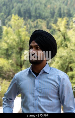 Ein Porträt bis zur Taille eines indischen Sikh-Mannes, der einen traditionellen schwarzen Turban und ein hellblaues langärmeliges Hemd mit Nadelstreifen trägt und weg schaut. Stockfoto