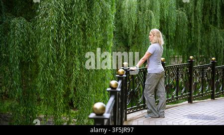 Eine mittelalte kaukasische Frau mit blondem lockigem Haar steht auf einem gepflasterten Gehweg in einem Park und lehnt sich an einen schwarzen verzierten Metallzaun mit goldener Kugel Stockfoto
