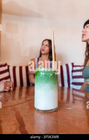 Nahaufnahme eines geschichteten Matcha-Lattes mit einem Strohhalm auf einem Holztisch, während junge Frauenfreunde sich in einem gemütlichen Café-Innenraum entspannen und unterhalten. Stockfoto