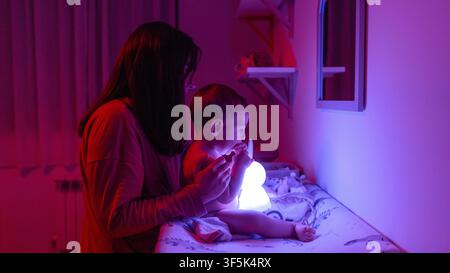 Mutter sitzt mit ihrem Baby auf einem Wickeltisch zu Hause und schaut auf ein leuchtendes Nachtlicht. Gemütliche Familien-Gute-Nacht-Routine in einem sanft beleuchteten Kinderzimmer, ruhig und s Stockfoto