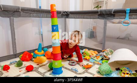 Neugieriges Kleinkind spielt in einem weichen Laufstall mit einem hohen Turm aus bunten Stapelbechern auf einer Matte, umgeben von Spielzeug in einem hellen, gemütlichen Raum. Stockfoto