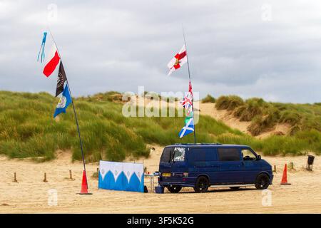 Ein Mann sitzt in einem Liegestuhl auf einem Sandstrand neben hohen Flaggenmasten, an denen eine Vielzahl von National- und Fußballflaggen weht. Southport, UK. Stockfoto