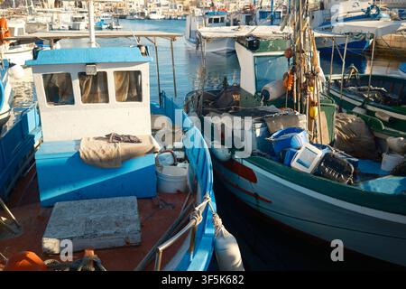 Reihen von verwitterten blauen, weißen, grünen und orangefarbenen Fischerbooten ruhen nebeneinander im sonnenbeschienenen Hafen von Favignana, Sizilien, mit Seilen, Netzen und ruhiger See Stockfoto