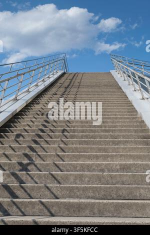 Eine breite Betonaußen Treppe führt aus einem niedrigen Blickwinkel steil nach oben direkt in einen hellblauen Sommerhimmel mit weißen Wolken. Stockfoto