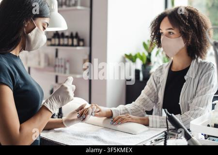 Neue Normalität und Kampf gegen das Virus im Schönheitssalon. Asiatische Meisterin in Schutzmaske arbeitet mit Nägeln und desinfiziert Hände mit Antiseptikum von Stockfoto