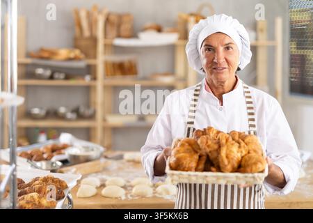 Reife lächelnde Bäckerin zeigt warme, leckere Croissants, die in einer kleinen Bäckerei gebacken wurden Stockfoto