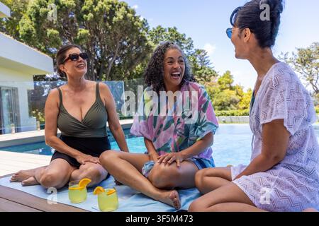 Verschiedene Freundinnen, die auf einem Handtuch am Pool sitzen, Zitrusgetränke schlürfen und Sonnenbrillen tragen. Hinterhof, Außenbereich, Sommer, Lebensstil, ungezwungen, sozial, Gespräch Stockfoto