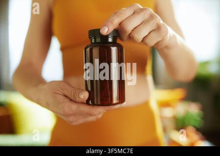 Eine Frau in einem orangefarbenen Oberteil hält eine dunkelbraune Plastikflasche und präsentiert ein Gesundheitsprodukt oder Vitaminpräparat als Teil eines Wellness-Lebensstils. Stockfoto