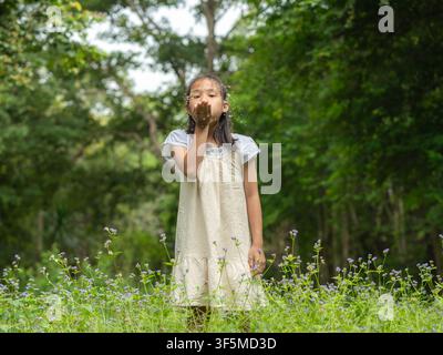 Asiatisches Mädchen in weißem Kleid, das Blumenblätter von ihrer Hand wegbläst und in einem Wildblumenfeld steht Stockfoto