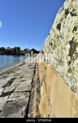 Eine Nahaufnahme-Perspektive einer verwitterten Steinmauer eines Reservoirs, die entlang ruhigen Wassers unter einem strahlend blauen Himmel verläuft, betont historische Mauerarbeit. Stockfoto