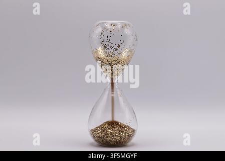 Eine elegante Glas-Sanduhr gefüllt mit glänzendem goldenem Glitzer, die auf einer weißen Oberfläche steht und den Wert der Zeit symbolisiert. Stockfoto