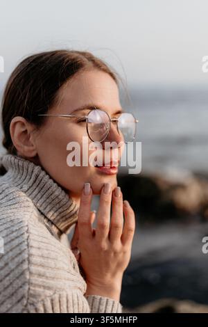 Frau mit Brille betrachtet junge Frau in einem beigen Strickpullover, die während einer friedlichen goldenen Stunde beim Sonnenuntergang auf den Ozean blickt Stockfoto