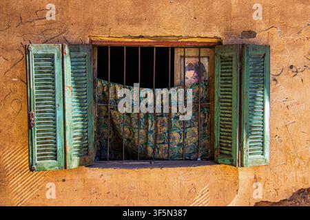 Ein verwittertes Fenster mit abgeblätterten grünen Holzläden und eisernen Sicherheitsgittern in Luxor, Ägypten, fotografiert am 16. Februar 2024. Ein gemusterter Vorhang pa Stockfoto