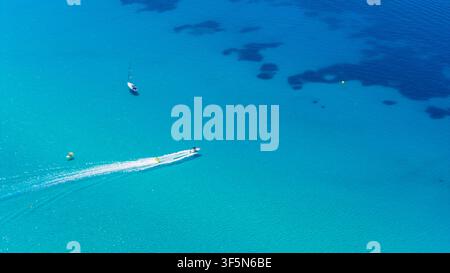 Klares blaues Wasser umgibt ein schnell fahrendes weißes Boot und ein kleines Segelboot, während dunkle Unterwasserflecken Kontrast über die offene Meereslandschaft erzeugen Stockfoto