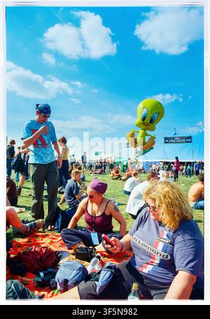 FESTIVALFREUNDE ENTSPANNEN IM SOMMERSONNENSCHEIN, READING FESTIVAL 1999, ROCKFANS UND MENSCHENMENGEN-SZENEN, READING, ENGLAND, 1999: Eine Gruppe von Freunden entspannt sich auf einer Decke in einem sonnigen Feld auf einem Musikfestival, eine Frau schaut auf ihr Handy, während ein großer gelber Tweety-Bird-Charakterballon in der Nähe schwebt. Foto: Rob Watkins. INFO: In den späten 1990er Jahren entwickelte sich das Reading Festival zum führenden alternativen Musikereignis Großbritanniens mit Rock, Indie, Punk, Metal, elektronischer Musik und aufstrebenden Künstlern. Seine vielfältige Besetzung zog riesige Menschenmengen an und half, die lebendige Festivalkultur dieser Ära zu prägen. Stockfoto