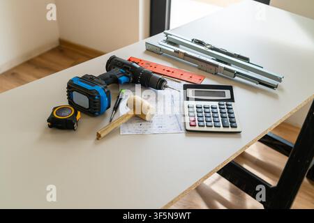 Bauwerkzeuge wie eine Akku-Bohrmaschine, ein Maßband und ein Taschenrechner werden an der Arbeitsstelle bewertet. Detaillierte Notizen und verschiedene Hardwarekomponenten sind ordentlich auf dem Tisch angeordnet. Stockfoto