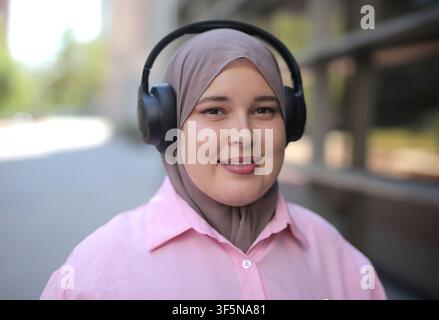 Junge Frau mit Hijab lächelt, während sie draußen Kopfhörer hört Stockfoto