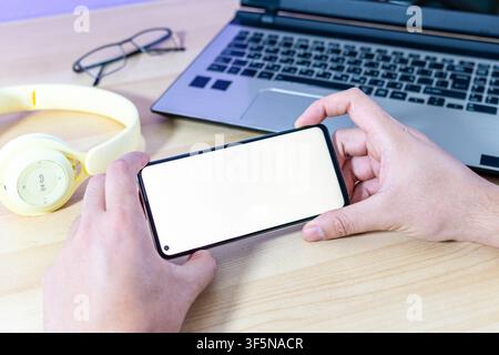 Zwei Hände halten ein Smartphone im Querformat über einem Schreibtisch mit gelben Kopfhörern und einem Laptop. Leerer Bildschirm für Mockups. Stockfoto