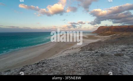 Weite Küstenansicht mit türkisfarbenem Meer, leerem Sandstrand und felsigen Hügeln unter weichen Wolken bei Sonnenuntergang. Friedliche Naturlandschaft mit offenem Raum und Tra Stockfoto