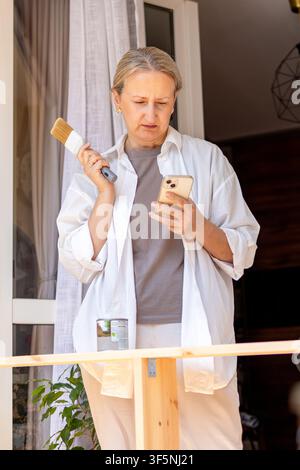 Frau hält einen Pinsel, während sie ein Tutorial auf dem Telefon überprüft. Konzept der Selbstbildung und des digitalen Lernens während eines DIY-Projekts. Stockfoto