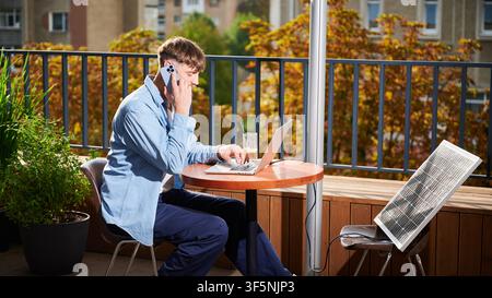 Man arbeitet mit Laptop und Smartphone am Tisch im Freien, mit einem Glas Eiskaffee. Solarpanel in der Nähe, der nachhaltigen, modernen, umweltfreundlichen Arbeitsbereich betont, der Technologie und Nachhaltigkeit vereint. Stockfoto