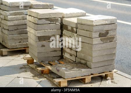 Betonplatten, die auf einer Holzpalette nahe einem städtischen Straßenbordstein gestapelt sind. Beton-Bordsteinfliesen in Stapeln für den Straßenbau und die Erhaltung von Pflasterflächen Stockfoto