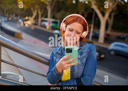 Rotschopf-Frau lächelt, hört Audio zu und benutzt ihr Telefon im Freien. Verbindung mit der Technologie in einer Stadtumgebung Stockfoto