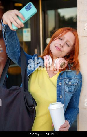 Rottbraune Frau posiert für ein Selfie mit einem Handy, hält eine Kaffeetasse und trägt Kopfhörer in einer städtischen Umgebung Stockfoto