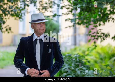 Ein älterer Mann, gekleidet in einem dunklen Anzug und einem weißen Hut, geht den Gartenweg entlang und strahlt Selbstvertrauen und Eleganz aus, während das Sonnenlicht durch die Bäume fällt Stockfoto