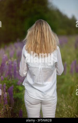 Eine Frau mit langen blonden Haaren schlendert durch ein lebendiges Feld mit lila Blumen und genießt einen schönen Sonnentag vor üppiger Grünflora im Hintergrund Stockfoto