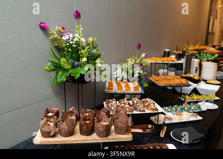 Eleganter Buffettisch zeigt verschiedene Gebäckstücke und frische Blumen. Schokoladenmuffins, Brezeln und Süßigkeiten sind ordentlich für die Gäste arrangiert. Bunte Flora Stockfoto